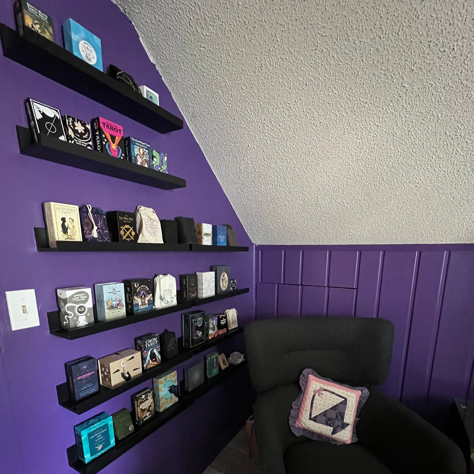The corner of a gable room with purple walls and a white popcorn ceiling. There is a grey chair in the corner with a purple, white, and pink pillow in it. On the wall are black shelves filled with tarot decks.